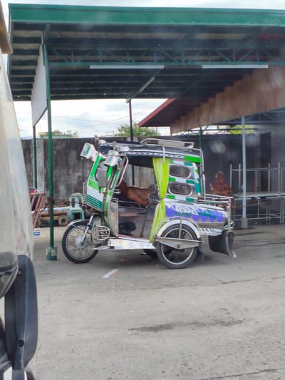 The image shows a colorful, decorated tricycle parked under a covered area. The tricycle is green and white with silver accents, and features a prominent green curtain. A man is seated inside the tricycle, partially visible, with his legs extended. In the background, another person is sitting outside the covered area, looking towards the right. The setting appears to be an outdoor market or a transportation hub in Cavite City, Philippines. The ground is paved, and a concrete wall runs along the back. There are metal racks and structures in the background, suggesting a commercial or service area. The lighting suggests it is daytime, with a cloudy sky. The overall impression is a snapshot of daily life in a busy, tropical location. No visible text is present in the image.