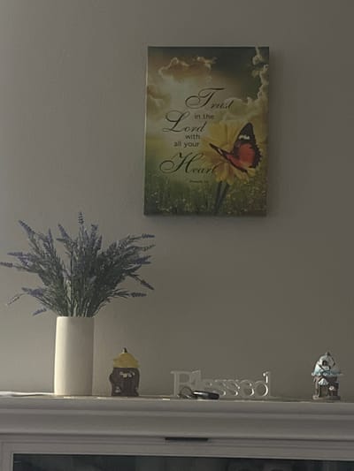 A still life photograph is shown of a mantelpiece with decorative items. On the left, a white vase holds a bouquet of artificial lavender. To the right of the vase, a small ceramic figurine resembling a bee with a yellow hat sits on the mantel. Further to the right, a decorative word art piece spells out "Blessed" in white, with a set of keys resting in front of it. On the far right, another small decorative figurine of a house with a blue roof is visible. Above the mantelpiece, hanging on the wall, is a framed inspirational artwork featuring text and a butterfly on a flower. The overall scene is static, with no people or discernible actions taking place. The environment appears to be an indoor domestic setting. The location is identified as Jacksonville, United States of America, though no specific city landmarks or context are provided to support this.