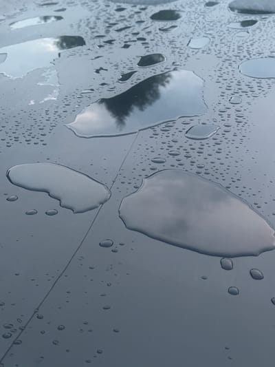 The image shows a close-up, high-angle view of a dark, smooth, flat surface covered in water droplets and several larger puddles. The surface appears to be outdoors, with reflections of a cloudy sky and possibly trees visible in the larger puddles. There are no people, animals, or specific landmarks visible that would indicate a location within the city of Lapės, Lithuania, or any other specific urban setting. The scene is purely naturalistic, focusing on the abstract patterns created by the water on the surface. The surface itself is segmented by a subtle, straight line running diagonally through the image, suggesting it might be a tiled or paneled structure.