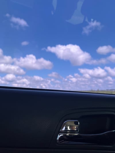 The image is taken from inside a vehicle, looking out through the window at a bright blue sky with scattered white cumulus clouds. A portion of the vehicle's interior is visible in the foreground, including a black textured armrest and a chrome-plated door handle. The reflection on the chrome handle shows a person's face. The background appears to be a slightly blurred landscape with a faint horizon line. The image was captured in the city of Марьевка, Russia.
