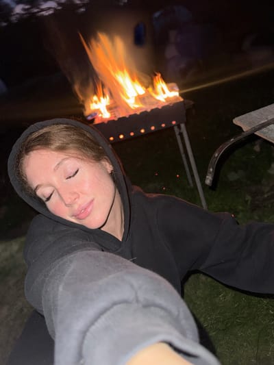 A young woman with reddish-brown hair and closed eyes is captured in a close-up selfie. She is wearing a black hoodie, with the hood pulled up over her head. Her eyes are shut, and her face has a serene expression, with her head tilted slightly. In the background, a bonfire burns brightly in a metal grill, casting an orange glow on the scene. The environment appears to be outdoors at night, with grass visible on the ground and hints of other people and objects in the dark periphery. The location is identified as Поварёнки, Russia.