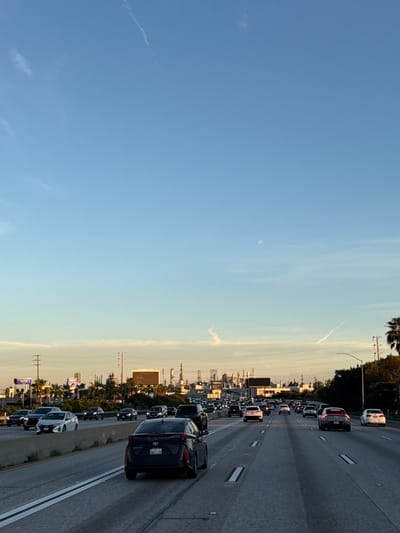 A congested highway is shown in this landscape. Numerous cars are traveling on the multiple lanes of the road, creating a traffic jam. Palm trees line the side of the road, suggesting a warm climate. In the distance, industrial structures like cranes and storage tanks are visible, indicating the presence of a port or industrial area. The clear blue sky above suggests fair weather.