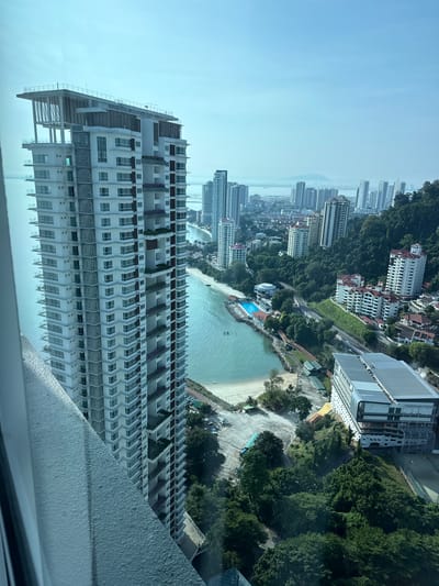 This is a high-angle, full shot of a coastal city skyline, likely George Town, Malaysia, viewed from a high-rise building. In the foreground, a tall, modern apartment building with white and grey balconies dominates the left side of the frame. The building features a unique architectural design at its top with open-air levels and a flat roof. To the right, the city unfolds with a crescent-shaped sandy beach bordering a turquoise body of water. Several other high-rise residential buildings are scattered along the coastline and further inland, with some nestled against a lush green hillside. A swimming pool with blue water is visible near the beach. Trees and vegetation cover the hillside and parts of the urban landscape. The sky is clear and bright, with a slight haze in the distance where more buildings and landforms are visible. The overall scene depicts a well-developed coastal urban environment with a mix of residential high-rises, natural greenery, and beachfront amenities.