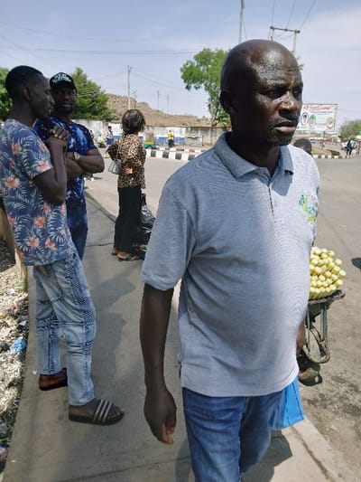 An outdoor scene in Communauté rurale de Payar, Senegal, depicts a street environment under a clear, bright sky. In the foreground, a dark-skinned adult male with a bald head and short facial hair is seen from the chest up, facing slightly to the right of the camera's perspective. He wears a light grey polo shirt with a green and white logo on the right chest and blue jeans. A blue plastic bag or item is held in his right hand. Adjacent to him on the right, a two-wheeled street vendor's cart holds a tall pile of yellow, cylindrical items, possibly sugar cane sticks.
To the left, two dark-skinned adult males are engaged in conversation on a concrete sidewalk. The man on the far left wears a blue floral short-sleeved shirt, light blue ripped jeans, and dark striped slide sandals, with his hands clasped. The man next to him wears a dark tie-dye patterned short-sleeved shirt, a dark baseball cap with a light emblem, and has his arms crossed. A watch is visible on his left wrist. Further back, a dark-haired woman is observed from behind, wearing a leopard-print top and dark trousers, carrying a white shoulder bag, with a large black bag at her feet.
The background features a paved road with a black and white striped curb. Across the street, various structures are visible, including one with a partially visible "WELCOME" sign featuring colorful graphics. Beyond these structures, a rocky, arid hill or small mountain rises. Sparse green trees and overhead power lines with utility poles are also present. The sidewalk in the immediate foreground exhibits minor debris.