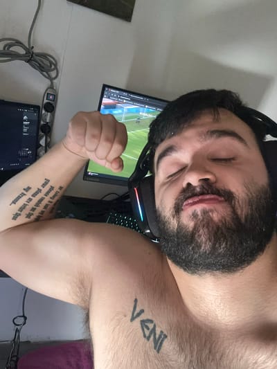 A man with a beard and closed eyes is wearing a headset and is situated in front of a computer setup consisting of two monitors and a keyboard. He has tattoos on his left arm and chest. No information suggests this photo was taken in Savigny-en-Véron, France.