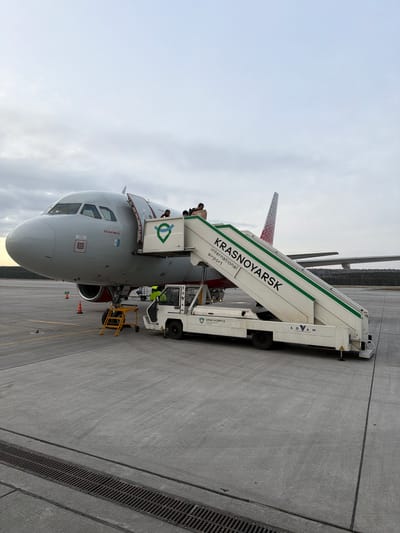 An airport scene in Емельяново, Russia, features an aircraft with a boarding stair vehicle connected to its front entrance. The plane's nose is prominently visible, exhibiting a gray-white color scheme with red accents near the engine area and tail. “Krasnoyarsk International Airport” is labeled on the boarding stairs. People are visible on the aircraft entrance with the boarding stairs. A smaller yellow utility vehicle is parked beside the stairs. The tarmac is gray concrete, and the sky is overcast.