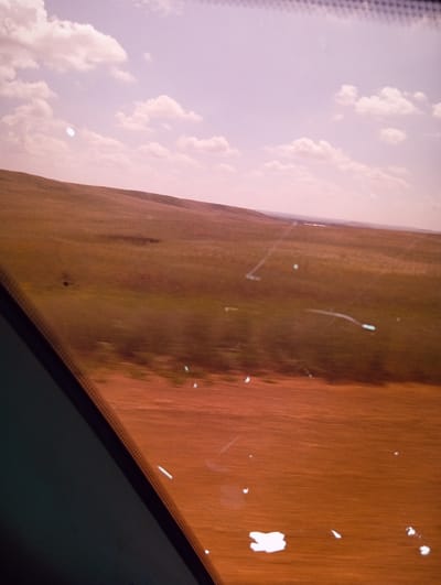 The image shows a view from a moving vehicle, looking out at a landscape under a hazy, pink-tinged sky with scattered clouds. The foreground is dominated by a dark, angular object, likely a window frame or pillar, with a textured edge. Beyond this, the landscape consists of rolling hills covered in dry, brownish-green grass. There are streaks and smudges on the window, creating distortions and artifacts in the image, particularly appearing as white or light blue smudges and lines across the landscape. In the distance, a very faint, linear arrangement of lighter colored objects is visible, possibly indicating structures or a road. The scene appears to be an open, rural area. The location of Aleksandrovka, Kazakhstan, is not depicted or suggested by any visual elements in the image.
