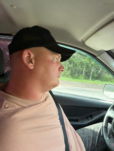 A man wearing a black baseball cap and a light pink t-shirt is seated in the driver's seat of a vehicle. A seatbelt is fastened across his chest. He appears to be resting or sleeping. A steering wheel is visible in the upper right corner. The car's window shows a view of trees and a road. The interior of the car is visible, including the dashboard and door panel. The location is not in Minderevo, Russia, as the scene does not exhibit any specific characteristics indicating its whereabouts.