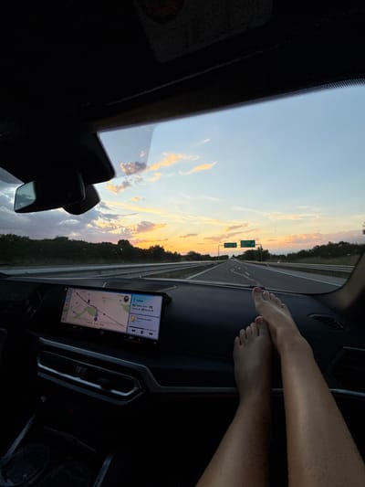 Here is a description of the image:
The image shows a view from inside a car driving on a highway at sunset. Feet are resting on the dashboard. A navigation system displays a map with a route marked to Sofia, Bulgaria from Cherna Gora, Bulgaria. Information indicates the route is 87km long and the ETA is 21:27.