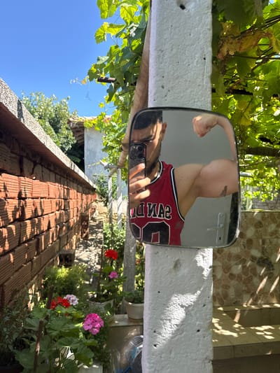 A man in a red tank top is taking a mirror selfie in a rural outdoor setting. He is flexing his bicep, and his face is partially visible in the mirror. The background shows a brick wall, green foliage, and a clear blue sky, suggesting a sunny day. The environment appears to be a yard or garden area. There is no indication of the city of Konop, Bulgaria.