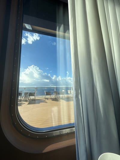 The image displays an interior perspective through a large, rounded rectangular window, framed in dark brown with visible metal rivets. A light-colored, vertically pleated curtain partially covers the right side of the view. Outside the window, a light brown wooden deck is visible, furnished with several unoccupied circular tables and chairs. Beyond the deck, a transparent railing precedes an expansive blue ocean. The sky above is clear blue, containing numerous white cumulus clouds. Direct sunlight illuminates the scene, creating reflections on the window glass. This environment is consistent with a coastal or maritime setting in the vicinity of Rum Cay, Bahamas. No individuals are present within the frame.