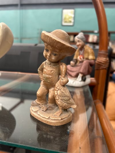 A medium-shot of a ceramic figurine of a boy with a hat carrying a goose. The figurine is light brown and stands on a decorated, circular base. In the background, out of focus, are additional figurines on a glass surface, with a wicker structure visible above. The scene is located within a room, as suggested by a light blue-green wall and window frame visible in the background. The image shows the scene on a piece of glass, with objects appearing on it. I cannot determine the location as Veracruz City, Mexico, as there are no indications in the image.