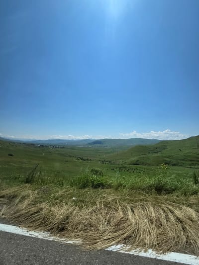 A panoramic, vertical view captures a vast, verdant landscape under a clear, bright blue sky, with the sun high and slightly to the left. The foreground is dominated by a strip of grey asphalt road on the left, marked with a white line. Adjacent to the road, a dense patch of dry, golden grass is prominent.
Beyond the road and grass, the scene opens up to rolling green hills that recede into the distance. The hills are covered in lush green grass, with patches of darker green suggesting trees or denser vegetation. In the mid-ground, a cluster of buildings, likely a village, is visible nestled within the valley. The architecture appears to be traditional, with red-tiled roofs. A few scattered trees are interspersed throughout the landscape.
Further in the distance, more rolling hills and mountains form the horizon, fading into a hazy, lighter blue. A few scattered white clouds are present in the sky, particularly towards the horizon. The overall impression is one of a peaceful, rural setting, likely in Khnkoyan, Armenia, given the context. The image appears to be taken during daylight, under sunny conditions. There are no people or animals clearly discernible in the scene.