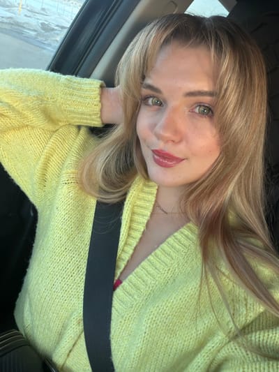 A young adult female, with long blonde hair and bangs, light skin, green eyes, and light makeup including reddish-pink lip gloss, is depicted from the chest up, positioned inside a vehicle, purportedly in the city of Азьмушкино, Russia. She wears a pale yellow, loose-knit V-neck cardigan over a visible pink undergarment and a black seatbelt diagonally across her chest. A delicate gold-colored necklace with a small pendant is visible. Her left arm is bent, with her hand resting behind her head. She is looking directly towards the viewer with a slight smile. The vehicle's door frame is partially visible on the left. Through the blurred car window, a bright outdoor scene with light-colored ground, possibly snow or ice, and indistinct distant elements suggests a winter or early spring environment during daylight hours.