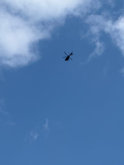 A medium-shot photograph captures the silhouette of a helicopter flying against a bright blue sky, partially covered by white clouds. The helicopter, positioned slightly to the left of the image's center, is dark and underexposed, indicating it is seen against a bright light source, likely the sun. Its main rotor blades are visible, along with the tail boom and tail rotor.

The sky is a vibrant blue, suggesting clear weather conditions, though interspersed with varying densities of white clouds. Some clouds are thin and wispy, while others form larger, puffier masses, particularly in the lower left and upper left quadrants of the frame. No specific activity beyond the helicopter's flight is discernible.

The time of day appears to be daytime due to the clear visibility and brightness of the sky. No people or visible text are present in the image. The scene conveys a sense of open space and aerial movement. The location context provided is Mont-Saint-Martin, France, though no specific visual cues in the image confirm this location.
