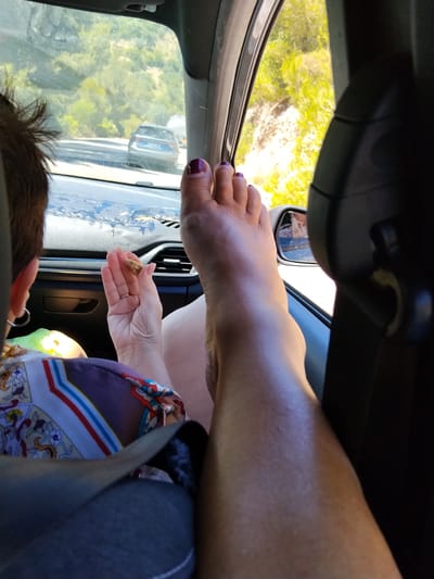 The image shows a person's bare foot, with toenails painted dark purple, resting on the dashboard of a car. The foot is positioned in the foreground, with the leg extending upwards. Through the car window, a hilly, green landscape is visible, with trees and a road where two cars are driving. The interior of the car, including the dashboard, seats, and a side mirror, is also visible. A person's hand, wearing a ring, is holding a small object, possibly food. The scene suggests a journey through a rural area in Mattinata, Italy, on a sunny day.