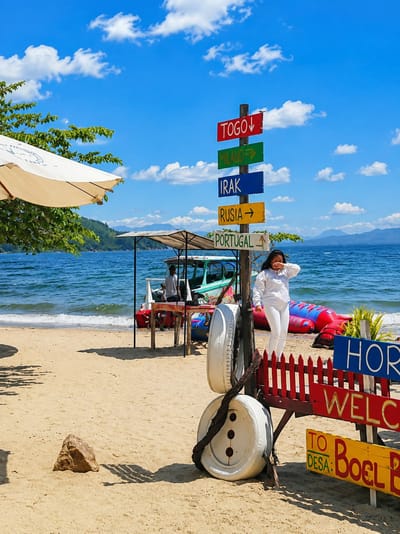In Lumban Bul Bul, Indonesia, this photograph captures a beach scene under a bright, cloudy sky. A directional signpost stands prominently, displaying destinations such as Togo, Milano, Irak, Rusia, and Portugal. To the right of the signpost, a person dressed in white stands near a vibrant red and blue inflatable water toy. In the background, the blue ocean meets the sandy shore, with a boat and a covered stall situated on the beach. The foreground features sand, a rock, and a decorative arrangement of tires and rope.