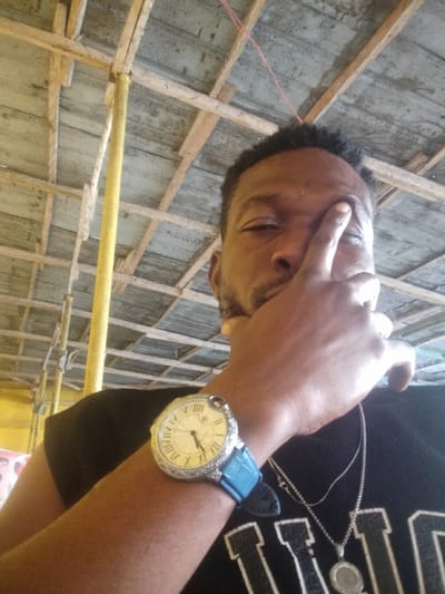 A man is seen in a selfie, with his right hand obscuring part of his face. He wears a black t-shirt with white lettering, along with two silver necklaces. A large watch with a blue band adorns his left wrist. The backdrop shows a wooden structure, possibly a ceiling or scaffolding, supported by yellow poles. The environment suggests he is either indoors or in a partially covered outdoor setting.