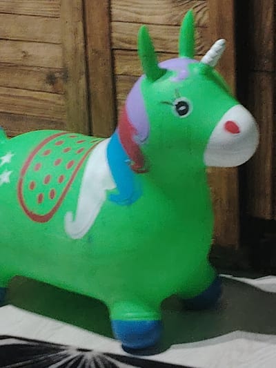 A bright green inflatable unicorn toy is positioned in the foreground of the image, its body detailed with red dots on a saddle-like area, white wings, and a blue mane. The unicorn has a white horn and a red nose. The background consists of a wooden-paneled wall. The toy is on a surface that appears to be a striped rug or mat. The image is cropped, focusing on the unicorn. There is no indication of the city of Prokhladny, Russia, or any specific location within it.