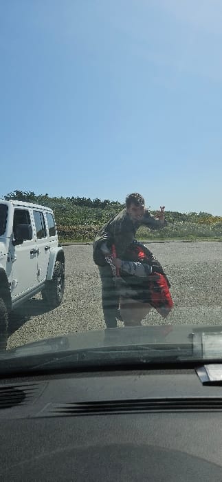 A white Jeep Wrangler is parked in a paved parking lot in Ocean Shores, Washington. The photo was taken from inside the vehicle, looking out the windshield. Two people are standing outside the vehicle. The person closest to the camera is a man in a gray hooded sweatshirt, with his back to the camera, and is holding a large red and black object that appears to be a life jacket. The man is smiling and has his right arm extended to the right side of the photo, with two fingers raised as if giving a peace sign. The second person is visible just behind the man, also wearing a gray hooded sweatshirt and jeans. The second person is facing forward, with his head turned slightly to the left. The background is a clear blue sky with a small amount of cloud cover. There are green bushes and trees in the background. The parking lot is gravel and the surrounding landscape is grassy and green. The photo is well-lit and has a bright, sunny appearance. The overall scene appears to be a casual, outdoorsy activity, likely related to a recreational activity. The photo was taken at the coordinates (latitude: 47.007, longitude: -124.168), which is a location in Ocean Shores, Washington, United States.