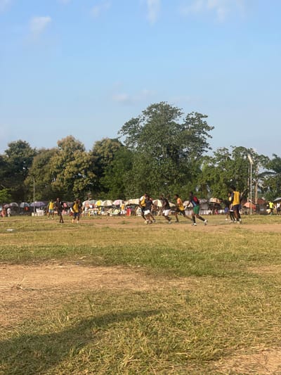 This is an outdoor, ground-level, wide shot of a soccer game being played on a dirt and grass field. The sky is a light blue with some scattered white clouds. In the background, there is a line of market stalls with umbrellas, and behind that, a dense line of green trees. Several players are visible on the field, actively engaged in the game. Most players are wearing casual athletic clothing, including t-shirts, shorts, and athletic shoes. Some are in yellow and black jerseys, while others wear various colors. A few spectators are visible standing near the market stalls. The field itself is uneven, with patches of dry grass and bare dirt. A single lamppost stands on the right side of the field.