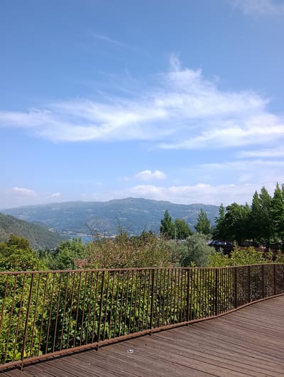 The image depicts a scenic viewpoint overlooking a valley with a body of water and rolling hills in the distance. In the foreground, there is a wooden walkway with a weathered metal railing. Lush green trees and bushes line the edge of the walkway and extend into the valley. The sky is a clear blue with scattered white clouds. The overall impression is one of a peaceful, natural landscape, possibly a park or a viewpoint in a mountainous region. The specific location in Terras de Bouro, Portugal, is suggested by the architecture and vegetation, characteristic of the region.