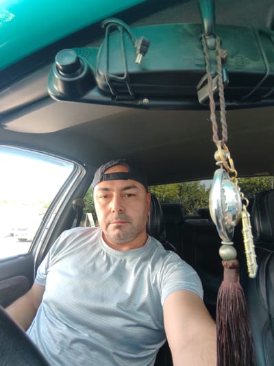 A man with a grey t-shirt and a black baseball cap is sitting in the driver's seat of a car. He has a serious expression on his face. The car's interior is visible, including the dashboard, rearview mirror with a dashcam attached, and a hanging ornament. Outside the car, trees and a bright sky can be seen, suggesting it is daytime. The image appears to be a selfie taken from inside the car.