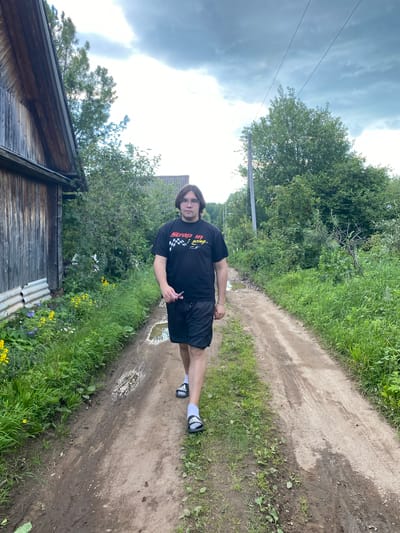 A person stands on a dirt path, with a wooden building to their left and lush greenery and trees on either side of the path. The person is wearing a black t-shirt with a checkered pattern and text, black shorts, and white slides with black stripes. The person is looking towards the camera with a neutral expression. The dirt path has tire tracks and puddles, suggesting recent rain. The sky is overcast with dramatic clouds, and the overall impression is of a rural or semi-rural setting in Novyi Ural, Russia. The time of day appears to be daytime, possibly late afternoon due to the lighting.