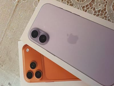 The image shows two iPhone boxes on a textured surface, possibly a tablecloth or placemat. The top box contains a light purple iPhone, and the bottom box contains an orange iPhone. Both phones are shown from the back, with their camera modules visible. The purple iPhone has two lenses, while the orange iPhone has three lenses. The Apple logo is visible on the back of the purple iPhone. The scene is illuminated by natural light, casting subtle shadows. There are no people or any other identifiable objects present in the image. The location in the city of Usoika, Bulgaria is not depicted or suggested by the image content.