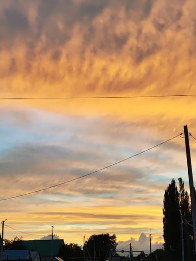 The image captures a dramatic sunset over the village of Novoursaevo, Russia. The sky is dominated by vibrant hues of orange and yellow, with streaks of pale blue visible in the lower portion. Wispy clouds are scattered across the sky, reflecting the warm light of the setting sun.

In the foreground, a dark silhouette of trees and the tops of buildings are visible against the colorful sky. A tall, thin utility pole with wires running across it is prominently featured on the right side of the frame, adding a stark contrast to the natural beauty of the sunset. Another utility pole and wires are visible on the left side of the image. The overall impression is one of a peaceful evening in a rural setting, with the elements of human infrastructure juxtaposed against the grandeur of the natural world.