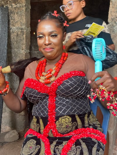 A woman adorned in traditional Igbo attire poses indoors. She wears a black and gold patterned dress edged with red beads, along with a layered coral necklace and hair ornaments. Another woman is seen partially in the image, fanning her with a small blue fan. The setting appears to be a textured concrete or stone structure. Overall, the scene captures a moment of traditional celebration or preparation.