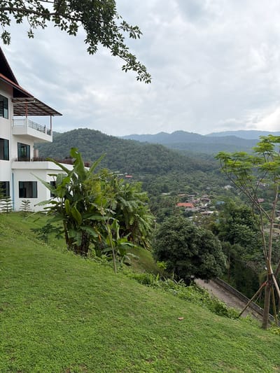 The image depicts a scenic overlook in the city of Ban Kao Dua, Thailand, characterized by lush greenery and a hazy sky. In the foreground, a steep grassy slope covered in small, dense green plants leads the eye towards the middle ground. Partially visible on the left is the exterior of a modern white building with balconies, featuring dark-framed windows and a dark-pitched roof. Adjacent to the building and spilling onto the slope are large banana trees and other tropical foliage. A paved pathway, bordered by a low concrete barrier and a young tree staked with poles, winds its way down the hillside on the right. The background reveals a dense, verdant valley filled with rolling, tree-covered hills, dotted with houses with red-tiled roofs, indicating a small settlement nestled within the landscape. The overall atmosphere is serene and natural, with the overcast sky suggesting a temperate day.