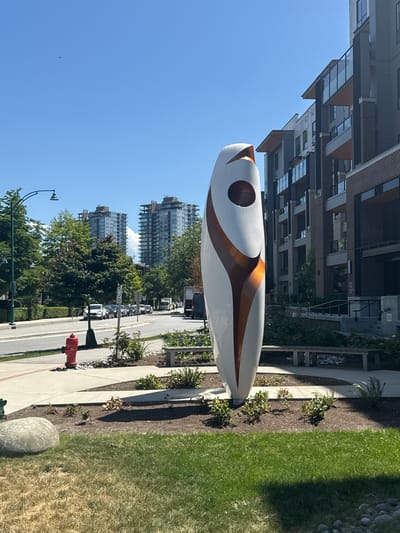 A vertical, white, oval-shaped sculpture with copper-colored accents stands in a park on a sunny day in Port Moody, Canada. The sculpture is positioned on a circular bed of mulch with small green plants. To the left of the sculpture, a red fire hydrant is visible on a paved sidewalk. Beyond the sidewalk is a street with parked cars and trees. In the background, a modern apartment building with balconies and glass windows is situated. Further in the distance, two tall residential buildings with reflective windows can be seen against a clear blue sky. A bird is flying in the sky.