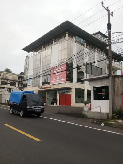 The image shows a black utility truck with a blue tarp driving on an asphalt road with a yellow dividing line. The truck is traveling in front of a modern, multi-story building. The building has large windows and appears to house commercial businesses, including "Daihatsu Astra Daihatsu" and "Hotel Algara," which is also indicated by a sign on the top of the building. There's a security or guard booth on the right side of the road near the building. Power lines are visible overhead. The location is most likely not in Taunggyi, Myanmar based on the commercial signs in the image. The scene takes place on a cloudy day.