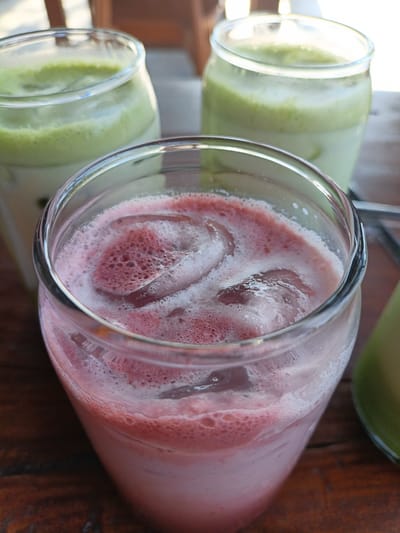 Three glasses of iced beverages are arranged on a wooden table. The glass in the foreground contains a pink beverage with foam and ice cubes, suggesting it might be a strawberry or raspberry drink. In the background, two glasses contain a light green beverage, possibly matcha or mint-flavored. The scene is outdoors, indicated by the natural lighting and the blurred background suggesting a cafe or restaurant setting. The wooden table surface has a visible grain. The city of Keudekruenggeukueh, Indonesia, is mentioned, implying this is a photograph taken within that location. There are no people visible in the image.