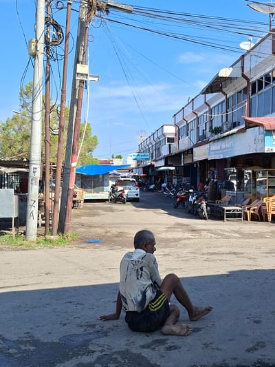 An elderly man with graying hair and a light-colored, patterned shirt sits on a paved road in Blangmetimu, Indonesia. His legs are crossed and his body is turned to the left, facing away from the camera. The man is wearing dark shorts with yellow stripes. In the background, there are several shops lining the street with awnings overhead. A white car and several motorcycles are parked in front of the shops. Utility poles with numerous wires are visible on the left side of the image, and trees can be seen behind them. The sky is clear and blue.