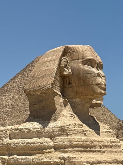 The image shows a close-up, low-angle view of a weathered stone carving against a clear blue sky. The carving appears to be a weathered section of an ancient sculpture, possibly a pharaoh's head, given the context of Giza, Egypt. The details of the carving are somewhat eroded but suggest a human face with distinct features. The stone itself is textured, with visible striations and rough surfaces, indicating age and exposure to the elements.

The lighting suggests it is daytime, with the sun casting shadows that highlight the three-dimensional qualities of the sculpture. The bright sky and strong sunlight are indicative of a clear, dry day, typical of the region. There is no visible text in the image. The focus is entirely on the texture and form of the ancient stonework, emphasizing its age and historical significance. The composition isolates the sculpted fragment from its surroundings, drawing the viewer's attention to the craftsmanship and the effects of time on the monument.