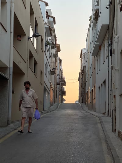 A man is walking down a narrow street in L'Escala, Spain. He is wearing a light-colored linen shirt and shorts, and carrying a purple bag in his right hand. The street is flanked by tall buildings with balconies and windows. The sky is a pale orange, suggesting it is either sunrise or sunset. The man appears relaxed as he walks. There is a sign on one of the buildings that is partially visible.