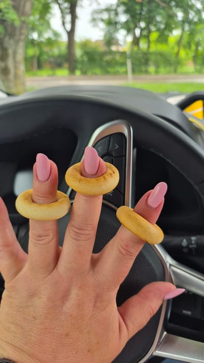 A close-up shot of a person's hand with three finger rings made from small, round, light brown bread rings, specifically bubliki, is visible. The rings are worn on the index, middle, and ring fingers. The fingers have neatly manicured, light pink almond-shaped nails. The hand is positioned in front of a dark-colored car steering wheel. The background is blurred, showing green trees and foliage, suggesting an outdoor setting. The image was captured in Jēkabpils, Latvia, likely while the person is inside a vehicle.