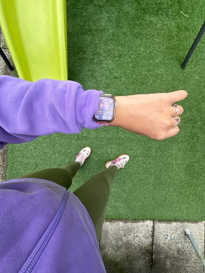 This image shows a person's legs and left arm, indicating they are standing. The person is wearing a purple hoodie and olive green leggings. On their left wrist, they are wearing a smartwatch that displays "08:31" and song information. The person is also wearing a white and floral-patterned athletic shoe. They are standing on a surface that appears to be artificial turf, suggesting an outdoor setting, possibly a park or a sports facility. A bright yellow object, possibly a bench or play equipment, is visible in the lower left corner. The ground next to the turf is a concrete or paved surface with some cracks and a small piece of debris. The overall impression is that the person is outdoors, perhaps on a break or before or after some physical activity, given the athletic attire and the time displayed on the watch. The weather appears overcast, as there are small droplets of water visible on the yellow object. The setting is likely a casual outdoor public space. No emotions are discernible as the person's face is not visible.