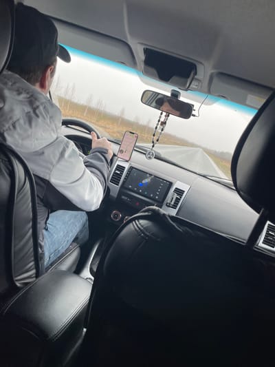 The image shows the interior of a vehicle, specifically the driver's seat and dashboard. A person wearing a black cap and a gray and white jacket is seated in the driver's seat with their hands on the steering wheel. A smartphone is mounted on the dashboard, displaying a navigation application. The car's infotainment system screen is visible between the air vents. A rearview mirror with a beaded ornament hanging from it is present. The background visible through the windshield suggests an overcast day in a rural area, possibly a road with trees on either side. Since Sidorovy Gory, Russia, is primarily a rural area, the scene likely depicts a local road.
