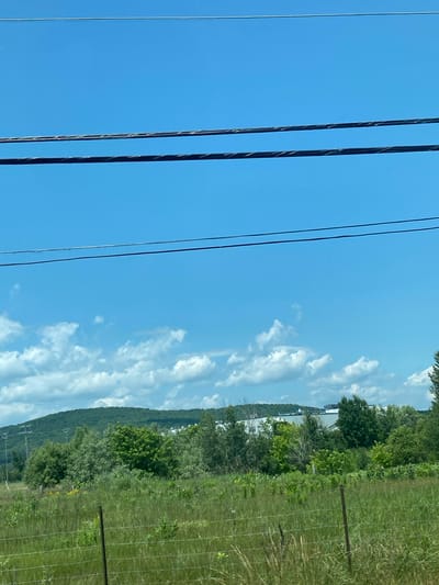 The image captures a rural landscape under a clear blue sky with scattered white clouds. In the foreground, a grassy field is partially obscured by a wire fence. Beyond the field, a dense line of trees and shrubs forms a green barrier, with a hilly, tree-covered landscape in the background. A white building or complex of buildings is visible on the crest of a hill in the distance, partially peeking through the trees. Several power lines stretch across the upper portion of the frame. The scene appears to be a natural setting in a rural area of Saint-André-Avellin, Canada, likely depicting a view from a road or elevated vantage point. No people or animals are visible in the frame.