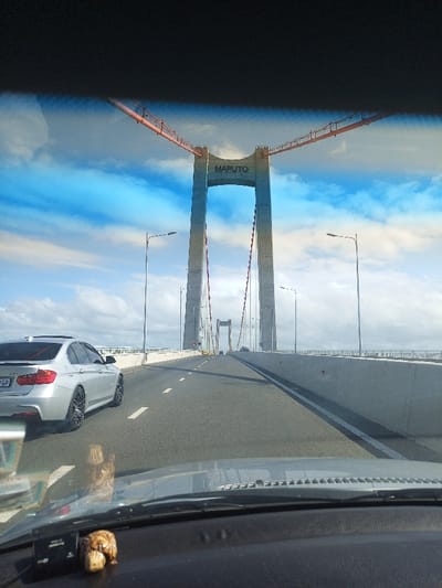 The image captures a front-facing view from inside a vehicle, showcasing the impressive Maputo-Katembe Bridge in Maputo, Mozambique. The perspective is from the driver's or front-seat passenger's position, with a dark dashboard and the lower part of the windshield visible in the foreground. A small, decorative item, possibly a carved figurine, rests on the dashboard.

The central focus of the photograph is the bridge's magnificent structure. Two towering concrete pylons stand prominently, with the word "MAPUTO" clearly inscribed near the top of the closest pylon. Thick, red-orange main cables stretch diagonally downwards from the top of the pylons, supporting the bridge deck via numerous vertical suspender cables, which are also visible. Another set of pylons can be seen in the distance, further along the bridge.

The sky above is a vibrant blue with scattered white clouds, indicating fair weather during daylight hours, likely morning or afternoon. The bridge features a wide, multi-lane roadway, with clear white lane markings. Concrete safety barriers line both sides of the bridge, and streetlights are spaced along its length.

In the left lane, a silver BMW sedan, possibly a 3-series, is seen driving ahead of the camera's vehicle. A portion of its license plate, which appears to include "GP," is visible on its rear, suggesting a South African registration. The road ahead appears clear, emphasizing the scale and modernity of the bridge and its surrounding environment.