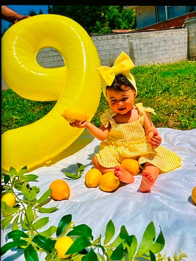 This is a photograph taken outdoors on a sunny day, likely in the Netherlands, given the location context of Nijmegen. The main subject is a baby, possibly celebrating a birthday, lying on a white sheet spread on green grass. The baby is wearing a yellow and white striped dress and a matching yellow bow on their head. The baby is smiling widely and appears happy and engaged.
Surrounding the baby are several yellow lemons, some on the sheet and one held by the baby. A large, inflatable yellow object, shaped like the number nine, is prominently featured in the foreground, partially obscuring the baby. To the right of the baby, there are green leaves and more lemons, suggesting a lemon-themed setup.
In the background, there is a hint of a brick building and more green foliage. The lighting suggests it is daytime, with clear, bright sunlight casting shadows. The overall scene is cheerful and festive, designed for a photoshoot. There is no visible text in the image.