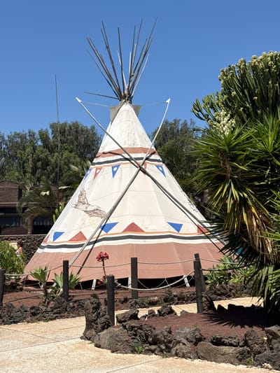 A teepee stands prominently in Tías, Spain, against a backdrop of lush greenery and a clear blue sky. The teepee is predominantly white with brown and red accents, featuring an eagle design on its front. It is surrounded by vegetation, including small trees and flowering plants. The base of the teepee is bordered by rocks and a paved walkway. A few dark wooden posts support the teepee's structure, adding to its rustic charm. The overall scene is sunny and appears to be outdoors, suggesting a warm climate.