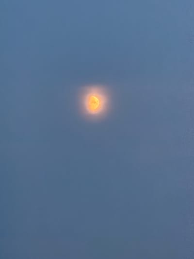 The image is a photograph of the moon, appearing partially obscured by thin clouds, in a dusky, blue sky. The moon itself is a bright, orange-yellow color and has a soft, diffused glow around it. The rest of the sky is a solid, muted blue, with a slight gradient of lighter blue at the top and darker blue towards the bottom. The image appears to be taken from an outdoor location in Kuseevo, Russia, though no specific landmarks or features of the city are visible. The composition is minimalist, focusing solely on the celestial body and the atmospheric conditions.