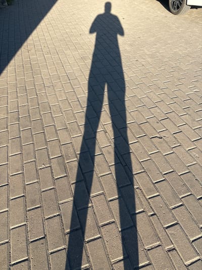 A long shadow of a person stands on a patterned paving stone surface. The shadow is cast by an unseen light source, likely the sun, creating a strong contrast between the illuminated and shadowed areas. The paving stones are rectangular and laid in a staggered pattern, with visible grout lines and some minor wear and tear on the surface. In the upper right corner, a portion of a car tire is visible, suggesting the setting is outdoors, possibly a street or parking area. The environment appears to be a sunny day, given the sharp shadow. There is no visible indication of the city of Suetovo, Russia, within the image itself.