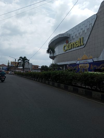 The image depicts a daytime urban street scene in Khajuripada, India, under an overcast sky. An asphalt road, appearing multi-lane, occupies the left and central foreground. A single individual, wearing a green garment and a helmet, rides a dark-colored motorcycle away from the viewer on the left side of the road. Further down the street, additional vehicles and various buildings, including one under construction, are visible. A tall street light pole stands on the left. The right side of the frame is dominated by a large, modern commercial building with a light-colored, textured facade and a curved architectural element. Prominent yellow signage on this building identifies it as "Citimall." Below the "Citimall" sign, a large blue and gold banner featuring ornate architectural designs and text, including the word "Dirgahayu," is displayed along the building's ground level. A dense row of green bushes, bordered by a curb with alternating black and white stripes, separates the road from the Citimall property. Several palm trees are present, and utility lines crisscross the sky.