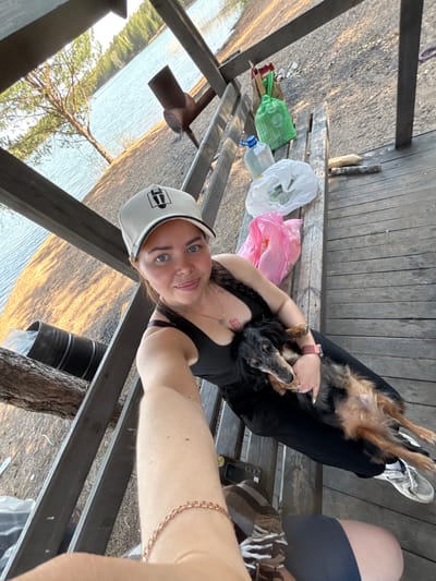 A rotated upward selfie photograph captures a woman and a dog on a wooden structure, possibly a deck or gazebo. The woman, wearing a black tank top and a white baseball cap, is holding the dog in her arms. The dog is small to medium-sized, with black and brown fur. The woman is also wearing a necklace, a bracelet, and sneakers.

Beside them, on the wooden surface, are various items, including plastic bags in white, green, and pink, and what appears to be a clear plastic bottle. The background shows a landscape with water and trees. The photograph seems to have been taken in Selychka, Russia, which appears in the geolocation data of the photograph.