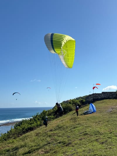 The image depicts a paragliding scene, likely taking place in Benoa, Indonesia, given the coastal setting. Several paragliders are visible in the sky against a clear blue background. One prominent green and white paraglider dominates the left side of the frame. Other paragliders appear smaller in the distance.

On the right side of the image, a grassy cliff overlooks the ocean. Several people are preparing for paragliding, with some already in flight. A person is standing near a blue paraglider, and another appears to be launching from the cliff edge. A red windsock is near a stone wall at the top of the cliff. The coastline below shows waves crashing against the shore, with trees and foliage lining the cliff's edge.